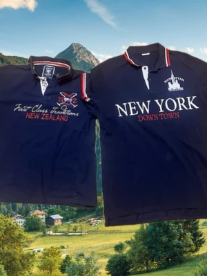  pike polo club  - Säljer Två stycken marinblå pikétröjor med krage och knappslå. Ena har broderat tryck 'NEW ZEALAND' och detaljer i vitt och rött, den andra har stort tryck 'NEW YORK DOWN TOWN' och Manhattan-motiv. Båda har sportig stil och röda/vita detaljer på kragen.