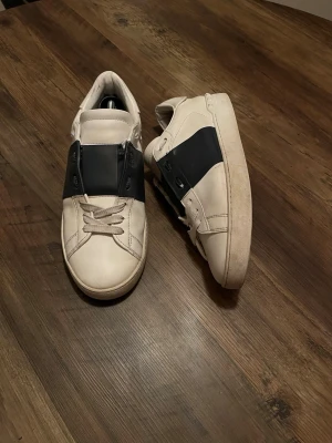 Vit svarta Valentino skor - Snygga vita sneakers med bred svart panel över snörningen. Skorna har en klassisk låg profil och är tillverkade i slätt lädermaterial. Perfekta för dig som gillar stilrena och moderna sneakers med en twist.