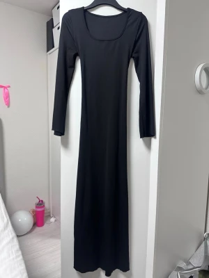 Svart långklänning med stretch bälte - En stilren svart långklänning med lång ärm och rundad urringning. Klänningen har en figurnära passform och är tillverkad i ett mjukt, stretchigt material som ger en bekväm känsla. Perfekt för dig som gillar minimalistisk och tidlös stil.