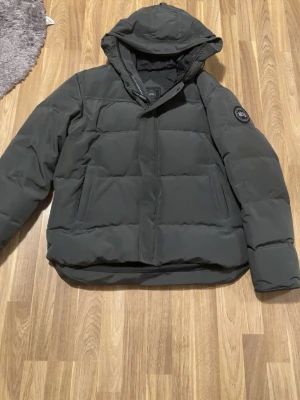 Canada Goose jacka - Säljer en svart pufferjacka från Canada Goose med huva och logotyp på ärmen. Jackan har quiltad design, två sidofickor och dragkedja med vindslå. Perfekt för kalla vinterdagar och riktigt varm tack vare det tjocka materialet. 