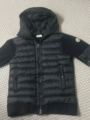 Svart dunväst med huva från Moncler - Snygg svart dunväst från Moncler med stickade ärmar och huva. Västen har två fickor med knappar och dragkedja framtill. Moncler-logga på ena ärmen och quiltad framsida i glansigt material. Perfekt för lager-på-lager och streetstyle.