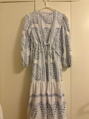 Vit klänning med blått blommönster - Lång vit klänning med blått broderat blommönster och lövdetaljer. Klänningen har puffärmar, v-ringning och knytband i midjan för en feminin siluett. Tillverkad i ett lätt tyg som ger en luftig känsla.