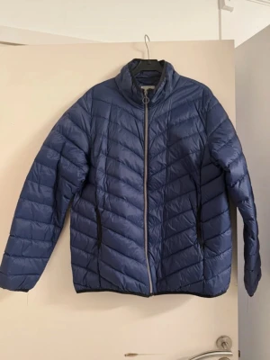 Mörkblå dunjacka med dragkedja - Snygg mörkblå dunjacka med hög krage och hel dragkedja framtill. Jackan har två sidofickor med dragkedja och ett quiltat mönster i diagonala linjer. Perfekt för kalla vinterdagar och ger en sportig look.