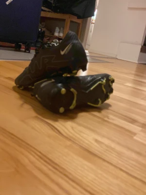Nike Mercurial svarta fotbollsskor - Säljer ett par svarta Nike Mercurial fotbollsskor med gula detaljer och dobbar. Skorna har en sportig design med tydliga Mercurial-loggor på hälen och Nike swoosh på sidan. Perfekta för dig som vill ha bra grepp och stil på planen.