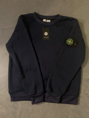 Mörkblå sweatshirt från Stone Island - Snygg mörkblå sweatshirt från Stone Island med klassisk rund hals och ikonisk logga på ärmen. Tröjan har ribbade muddar vid ärmslut och nederkant. Perfekt för en clean och stilren look.
