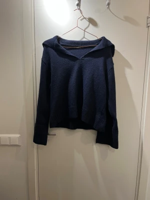 Mörkblå stickad hoodie - Mörkblå stickad med en krage och vida armar med en slits längst ner på armen, har knappt använt tröjan och tröjan är i bra skick inga hål eller skador.