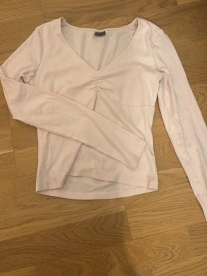 Ljusrosa långärmad topp med v-ringning - Söt ljusrosa topp med lång ärm och v-ringad hals. Toppen har en snygg rynkning framtill vid bysten och är gjord i mjukt bomullsmaterial. Perfekt att styla med jeans eller kjol för en enkel men trendig look.