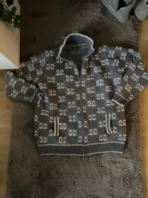 Grå ziptröja med mönster från G0cci - Grå stickad ziptröja från G9cci med vitt GG-mönster över hela tröjan. Tröjan har hög krage, dragkedja framtill och ribbade muddar samt fickor med vita detaljer. Perfekt för dig som gillar exklusiv streetstyle.passar L M