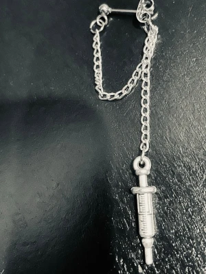 By prisförslag- 1 st Silvrigt örhänge med spruta och kedja - Unikt silvrigt örhänge med en detaljrik sprutberlock som hänger i en tunn kedja. Örhänget har en cool och edgy vibe, perfekt för dig som gillar statement-accessoarer. Materialet är metall i silverfärg och designen är inspirerad av medicinska teman.