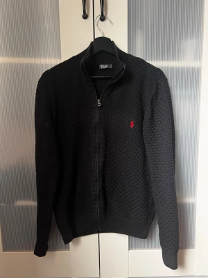 Polo Ralph Lauren Cardigan - Svart stickad tröja med dragkedja från Polo Ralph Lauren. Tröjan har ett diskret rutmönster, ribbade muddar och en liten röd logga broderad på bröstet. Hög krage och lång ärm för en clean och klassisk look. 
