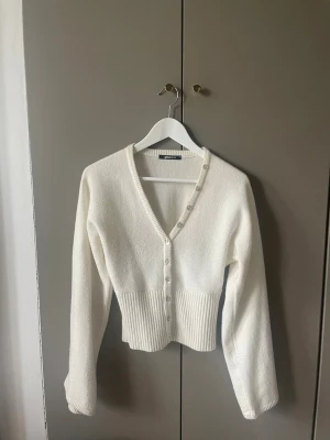 Vit ribbad kofta från Gina Tricot - Säljer en krämvit kofta från Gina Tricot med v-ringning och knappar framtill. Koftan har långa ärmar och bred ribbad midja som ger en snygg siluett. Perfekt att slänga över en topp för en clean look.