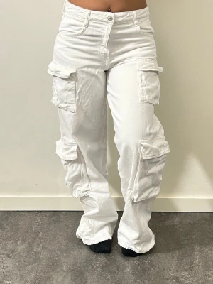 Vita cargobyxor med fickor - Säljer ett par vita cargobyxor med flera stora fickor på sidorna och bak. Byxorna har en avslappnad passform och raka ben. 