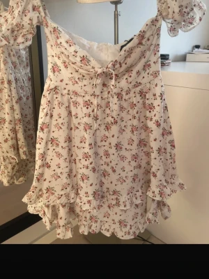 Vit blommig kortklänning med volang - Supersöt vit kortklänning med små blommor i rosa och grönt. Klänningen har volangkant nertill, knyt framtill och puffiga korta ärmar. Perfekt för sommarens alla tillfällen!