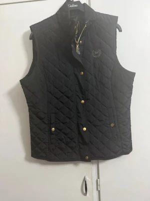 Svart quiltad dunväst från my wear - Snygg svart dunväst från  my wear med quiltat mönster och guldfärgade knappar. Västen har ståkrage, två fickor framtill och ett broderat emblem på bröstet. Perfekt för lager-på-lager och ger en stilren look.