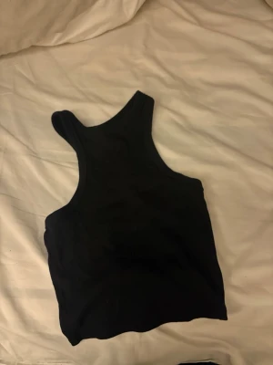Svart cropped ribbad tanktop - Svart cropped tanktop med ribbad struktur och brottarrygg. Perfekt för en avslappnad och trendig stil. Mjuk och stretchig känsla, passar till många olika outfits.