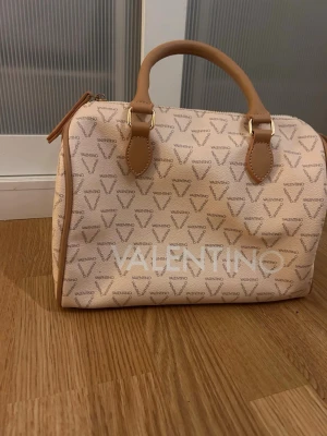Beige handväska från Valentino - Snygg beige handväska från Valentino med logomönster över hela väskan och stora vita bokstäver framtill. Väskan har två handtag i ljusbrunt konstläder och dragkedja upptill. Rymligt fack inuti och klassisk boxig form. Ny pris 1459kr 