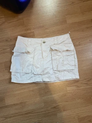 Vit kortkjol med stora fickor - Snygg vit kortkjol med två stora fickor framtill och knappstängning i midjan. Kjolen har bälteshällor och är gjord i ett lätt jeanstyg, perfekt för varma dagar. Enkel och stilren design som passar till många olika outfits.