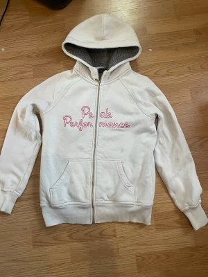Vit Peak Performance hoodie med teddyfoder - Säljer en vit hoodie från Peak Performance med dragkedja och rosa broderad text på bröstet. Hoodien har mysigt grått teddyfoder på insidan och en stor huva. Praktiska fickor framtill och ribbade muddar vid ärmar och nederkant. Perfekt för kalla dagar.