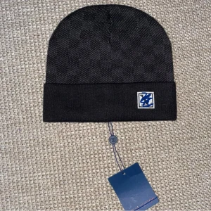 Svart l/v beanie - Snygg svart mössa från L/V med diskret rutmönster och vikt kant. På framsidan sitter en liten patch med blå L/V-logga. Klassisk passform och stilren design, perfekt för dig som gillar exklusiva accessoarer.