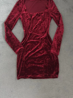 Vinröd sammet klänning långärmad - Supersnygg vinröd kort klänning i mjuk sammet med långa ärmar och fyrkantig ringning. Perfekt för dig som vill sticka ut med en elegant och trendig look. Klänningen har en kroppsnära passform och känns lyxig tack vare det glansiga materialet. Använt en gång, inga slitage!