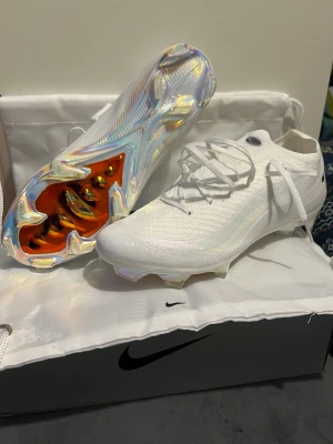 Vita Nike Mercurial fotbollsskor - Säljer ett par vita Nike Mercurial fotbollsskor med skinande, iriserande sula och klassisk låg profil. Skorna har snörning och en strömlinjeformad design i syntetmaterial för bästa grepp och känsla på planen. Perfekta för dig som vill sticka ut på matchen.