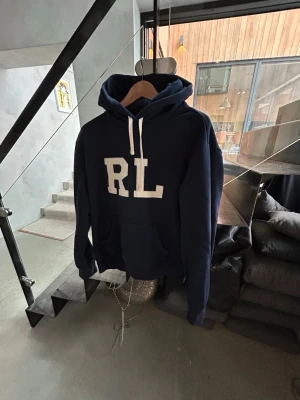 RL hoodie - Snygg mörkblå hoodie från Ralph Lauren med stora vita RL-bokstäver på bröstet. Hoodien har en klassisk huva med vita dragsnören och en magficka. Tillverkad i mjukt bomullsmaterial som är perfekt för chill dagar.