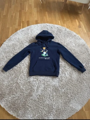  Polo Ralph Lauren Hoodie  - Mörkblå hoodie från Polo Ralph Lauren med den klassiska Polo Bear-trycket framtill. Hoodien har huva med dragsko och en stor magficka. Tillverkad i mjukt bomullsmaterial och har ribbade muddar vid ärmslut och nederkant. Perfekt för en avslappnad och cool stil.