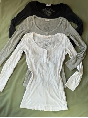  toppar från Marwin 99st - Tre långärmade toppar från Marwin i svart, grått och vitt. Alla har rund halsringning, knappar framtill och tryckt logga på bröstet. Slim fit-modell i mjukt material, perfekta för lager på lager eller som basplagg.