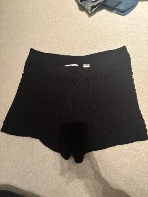 Svarta virkade shorts med snörning - Snygga svarta virkade shorts med snörning i midjan. De har en högmidjad passform och är stickade i ett luftigt mönster som ger en cool och avslappnad vibe. 