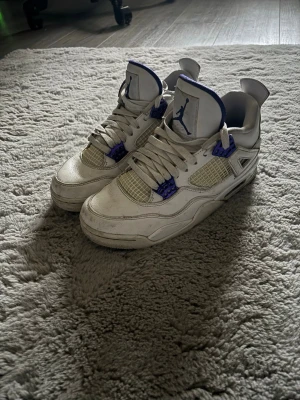 Air Jordan 4 vit/lila sneakers - Säljer ett par Air Jordan 4 sneakers i vitt med lila detaljer och meshpaneler. Skorna har klassisk hög siluett, vita skosnören och Jumpman-logga på plösen. Materialet är en mix av läder och textil, med coola plastdetaljer vid snörningen.