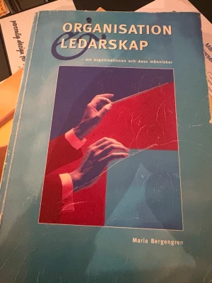 Organisation & Ledarskap bok - Kurslitteratur med titeln 'Organisation & Ledarskap' av Maria Bergengren. Boken har ett blått omslag med grafisk illustration av händer och dokument i rött och blått. Perfekt för dig som pluggar organisation och ledarskap.