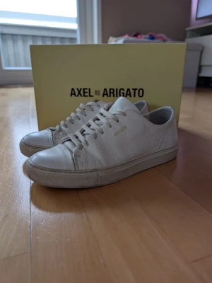 Vita Axel Arigato sneakers i läder - Stilrena vita sneakers från Axel Arigato i premiumläder med diskret guldfärgad logga på sidan. Skorna är i storleken 39 i fint skick det finns en liten repa på ovansidan av skon och vid loggan på höger skon! Limmet emot sulan har börjat släppa lite. 