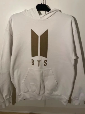 Vit BTS hoodie med logga - Vit hoodie med stor BTS-logga i brunt framtill. Klassisk känguruficka och huva. Mjuk och skön, perfekt för dig som älskar K-pop och vill visa ditt stöd för BTS. Ribbade muddar vid ärmslut och nederkant. Passar till chill dagar och konserter.