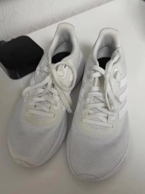 Vita Adidas sneakers med mesh - Snygga vita sneakers från Adidas med klassiska tre ränder på sidan. Skorna har en låg profil och är tillverkade i ett ventilerande meshmaterial med vita snören och sula. Perfekta för en clean och sportig look.Strl 40,5