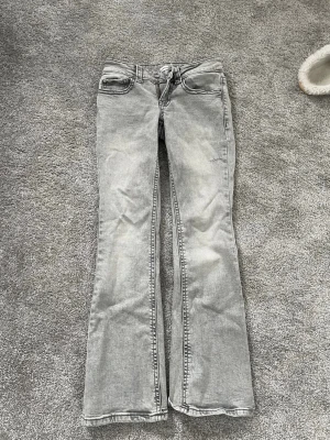Grå bootcut jeans med fickdetaljer - Säljer ett par lågmidjade ljusgrå bootcut jeans med slitna detaljer och knappfickor bak. Dom är i storlek 34 men dom är väldigt korta så skulle passa nån som är under 155 då de är väldigt små, mer som ett par jeans för ett barn.