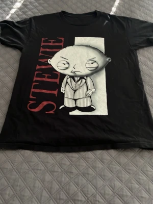 Svart Stewie t-shirt Family Guy - Cool svart t-shirt med stort tryck av Stewie från Family Guy i kostym på framsidan och texten 'STEWIE' i rött. T-shirten har rund hals och korta ärmar. Perfekt för dig som gillar tecknade serier och vill sticka ut med en unik print.