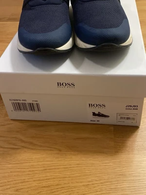 Marinblå sneakers från BOSS - Snygga slip on marinblå sneakers från BOSS med vit och svart sula. Ovandelen är i mesh och syntetmaterial för en sportig look. Skorna har en låg profil och klassisk design med diskreta detaljer. Perfekta för dig som gillar stilrena och bekväma sneakers.   Använda få gånger, bra skick. Originalkartong ingår men kvitto saknas. Storlek: 40 Nypris: ca 1 500 - 2 000. Mitt pris: 1 100 kr men kan diskuteras. Kontakta mig vid intresse. 