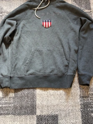 Grå hoodie från Gant med logga - Snygg grå hoodie från Gant med broderad logga och amerikanskt sköldmärke på bröstet. Klassisk känguruficka framtill och justerbar huva med vita snören. Tillverkad i mjukt bomullsmaterial som är skönt att ha på sig.