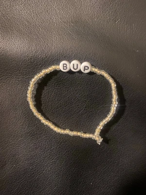 Pärlarmband med bokstäver BUP - Säljer ett handgjort armband med små genomskinliga pärlor och tre vita bokstavspärlor som stavar 'BUP'. Armbandet har en enkel och stilren design, perfekt för dig som gillar personliga accessoarer.