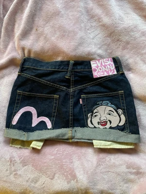 Evisu minikjol med tryck och brodyr - Storlek 28 men är klippt och kort så skulle säga storlek xs Unik mörkblå jeanskjol från Evisu med rå kant, rosa målat märke på ena bakfickan och broderad ansikte på den andra. Klassisk denim med kontrastsömmar och cool detalj med Evisu-logga upptill. Perfekt för dig som vill sticka ut. 
