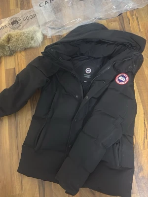 Canada goose Wyndham red badge/röd  - En jätte fin Canada goose jacka Wyndham är modellen jag har använd den ungefär 1 månad och den är jätte varm osånt ända skadan är att där inne i jackan på högra ficka så är hela fickan öppen alltså menat att på båda sidor finns det på högra sidan en ficka uppe o en ficka nere men den som splittrar så d blir e fickor är typ lite sönder bara lite lös fortfarande två fickor men trådar har lossnat och det finns mycket dun som är där inne helt normalt för en dun jacka som denna annars är den perfekt