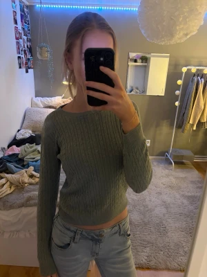 Grå-grön kabelstickad croptröja - Säljer en grå kabelstickad croptröja med lång ärm och ribbade muddar. Tröjan har ett klassiskt flätmönster och är tillverkad i mjukt stickat material. Perfekt för att skapa en trendig look med jeans eller kjol.