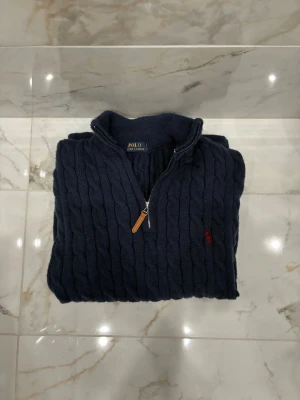 Mörkblå kabelstickad tröja Polo Ralph Lauren - Kabelstickad mörkblå tröja från Polo Ralph Lauren med halv dragkedja och ribbad krage. Tröjan har ett litet rött broderat logomärke på bröstet och är tillverkad i ett mjukt, tjockt material som passar perfekt för kyliga dagar.