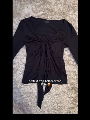 Svart omlottopp med knyt framtill - Trendig svart omlottopp från Gina Tricot med lång ärm och snygg knytning framtill. Djup v-ringning och figurnära passform som ger en stilren siluett. Perfekt att styla med jeans eller kjol för en cool look.