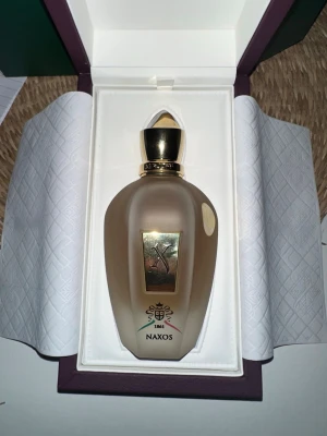 Xerjoff Naxos Eau de Parfum 100ml - Lyxig parfym från Xerjoff, Naxos, i en frostad glasflaska med gulddetaljer och elegant lock. Flaskan rymmer 100 ml och har exklusiv design med märkets logga och dekorativa inslag. Perfekt för dig som gillar unika och sofistikerade dofter. Använd en sprita bara väldigt lyxig för mig