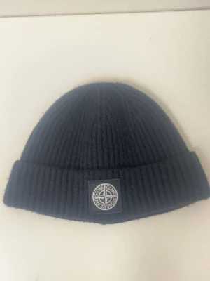Svart ribbstickad mössa Stone Island - Svart ribbstickad mössa från Stone Island med uppvikt kant och klassisk logopatch framtill. Snygg och enkel design som passar till det mesta. Materialet är mjukt och värmande, perfekt för kyliga dagar.