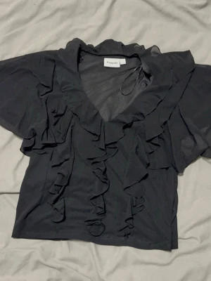 Svart volangblus från Kappahl - Snygg svart blus från Kappahl med v-ringning och stora volangdetaljer framtill och på ärmarna. Blusen har korta ärmar och är tillverkad i ett lätt, tunt material som ger ett luftigt intryck. Perfekt för dig som gillar en feminin och trendig look.