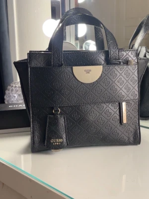 Svart handväska från Guess - Snygg svart handväska från Guess med präglat monogrammönster över hela väskan. Väskan har två handtag, guldfärgade detaljer och en tag med Guess 1981. Tillverkad i konstläder med stilren, fyrkantig form och logga framtill. Skriv för mer info mkt fin fortfarande 