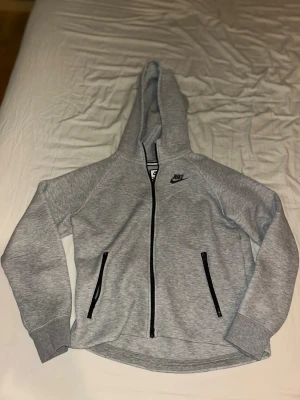 Grå Nike hoodie med dragkedja 156-166 - Säljer en grå tech fleece från Nike i storlek 156-166 . Hoodien har svart Nike-logga på bröstet, dragkedja framtill och två svarta dragkedjeförsedda fickor. Materialet är mjukt och bekvämt, perfekt för chill dagar. Huva och ribbade muddar på ärmarna. Den har bara varit använt typ 2-3 gånger den är köpt från Zalando