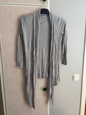 Ljusgrå lång kofta från KappAhl - Lång, tunn och ljusgrå kofta från KappAhl med öppen front och asymmetrisk nederkant. Koftan har långa ärmar och är tillverkad i ett mjukt, stretchigt material som faller snyggt. Perfekt att slänga över en t-shirt eller linne.